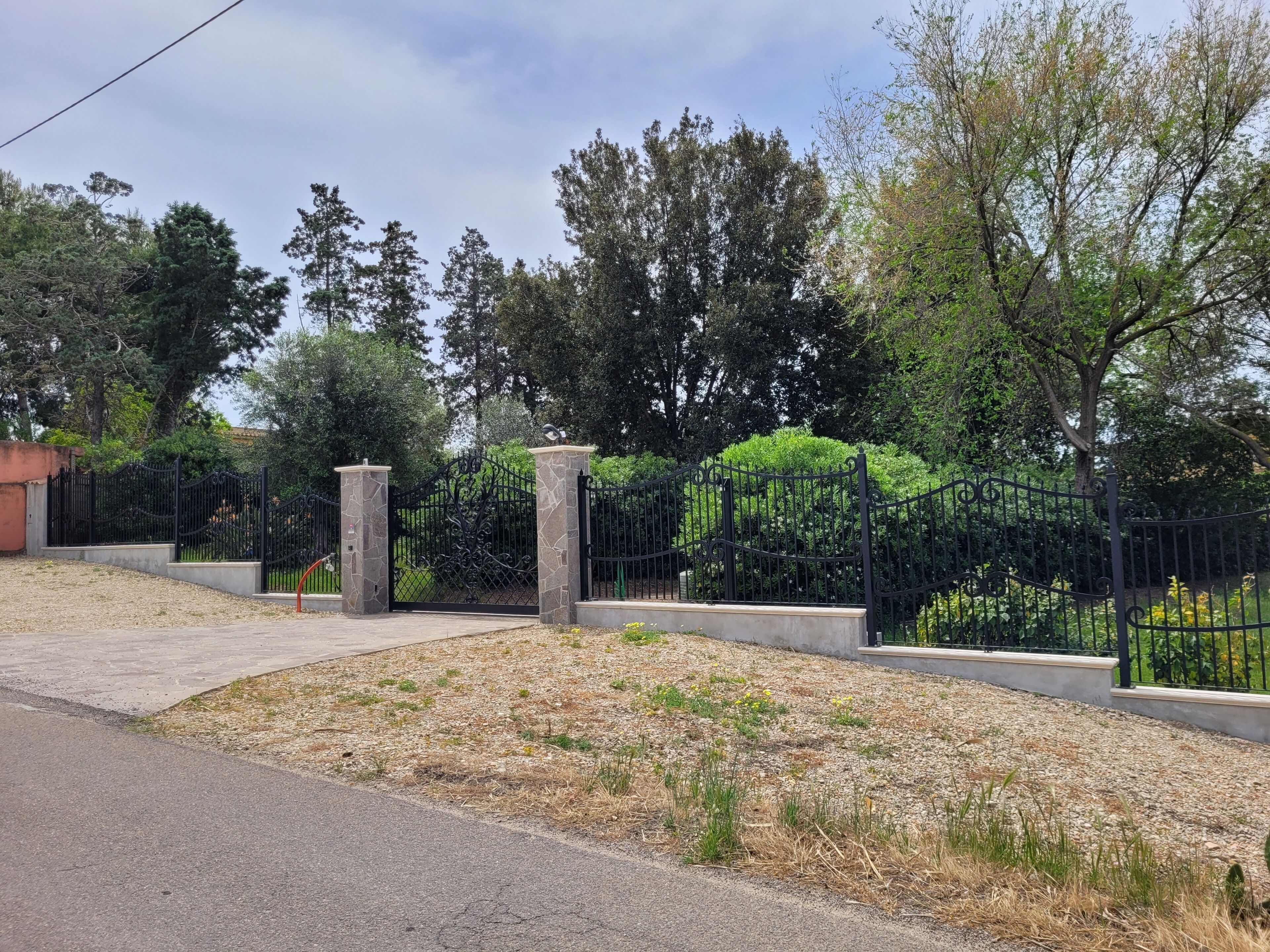 A gated entrance with a metal fence, stone pillars, and a pathway leading to a landscaped area with trees and shrubs.