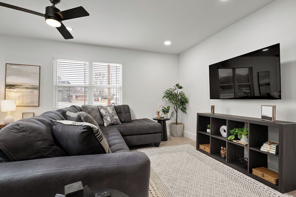 A living room with a sectional sofa, a TV mounted on the wall, a ceiling fan, and a window with blinds.