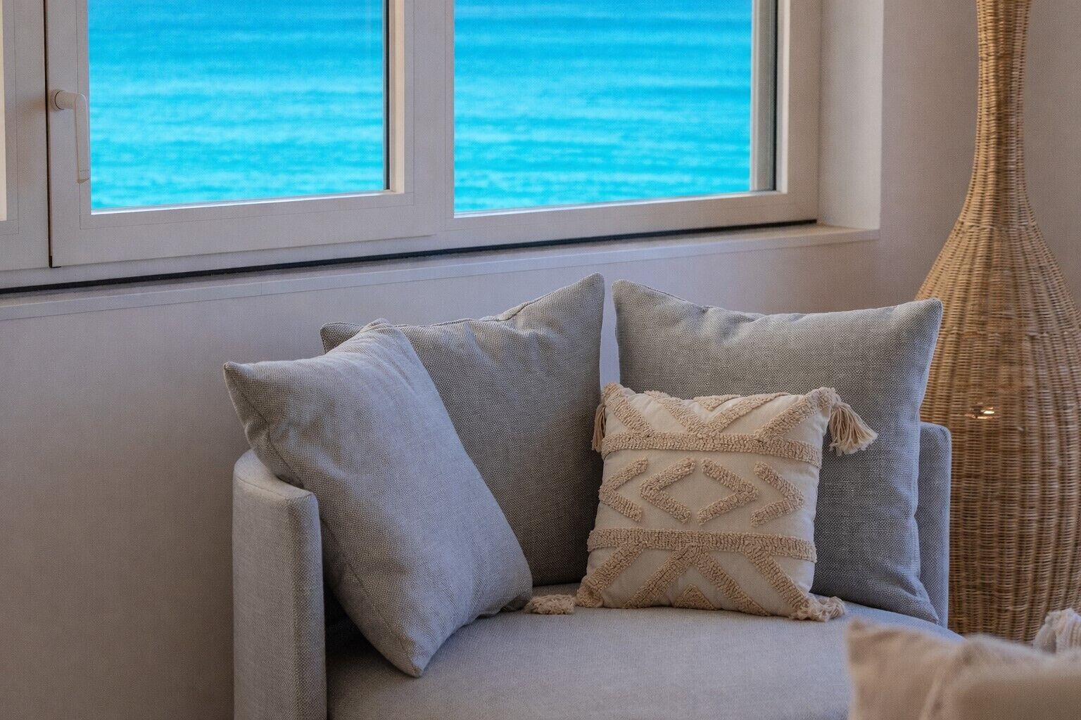 Ein Sofa mit Kissen, ein Korbstuhl und Blick auf das Meer durch ein Fenster.