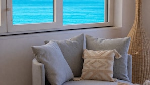 A sofa with cushions, a wicker chair, and a view of the ocean through a window. - Marzamemi on the beach SicilyMyHome (Marzamemi)