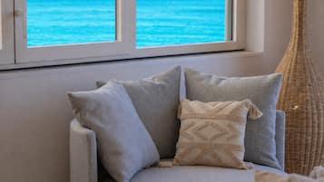 Ein Sofa mit Kissen, ein Korbstuhl und Blick auf das Meer durch ein Fenster.