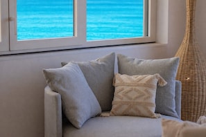 Ein Sofa mit Kissen, ein Korbstuhl und Blick auf das Meer durch ein Fenster.