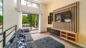 Ett modernt vardagsrum med en stor TV, en soffa och en balkong med utsikt över grönområden.
