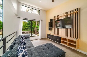 Ein modernes Wohnzimmer mit großem Fernseher, einer Couch und einem Balkon mit Blick auf Grünflächen.