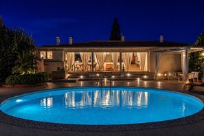 A swimming pool with a stone border, a covered outdoor seating area, and a building with a tiled roof in the background. - Villa Alexandra SicilyMyHome (Santa Maria del Focallo)