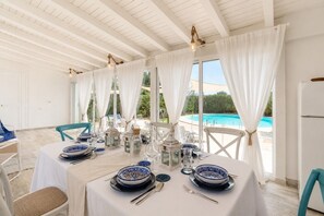 A dining area with a pool view, white curtains, and a table set with blue and white plates. - Villa Alexandra SicilyMyHome (Santa Maria del Focallo)