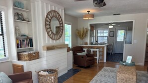 A spacious living area with a fireplace, a brown leather sofa, a wicker basket, and a kitchen in the background. - Steps to Beach - Prime Location Beach Bungalow – Beach Gear (Holmes Beach)