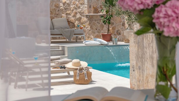 Area piscina con muretto in pietra, sedie a sdraio, un tavolino con cappello e cesto, e un libro aperto.
