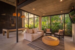 Ein modernes Wohnzimmer mit Poolbillard, einer Couch, Korbstühlen und großen Fenstern mit Blick auf einen üppigen Garten.