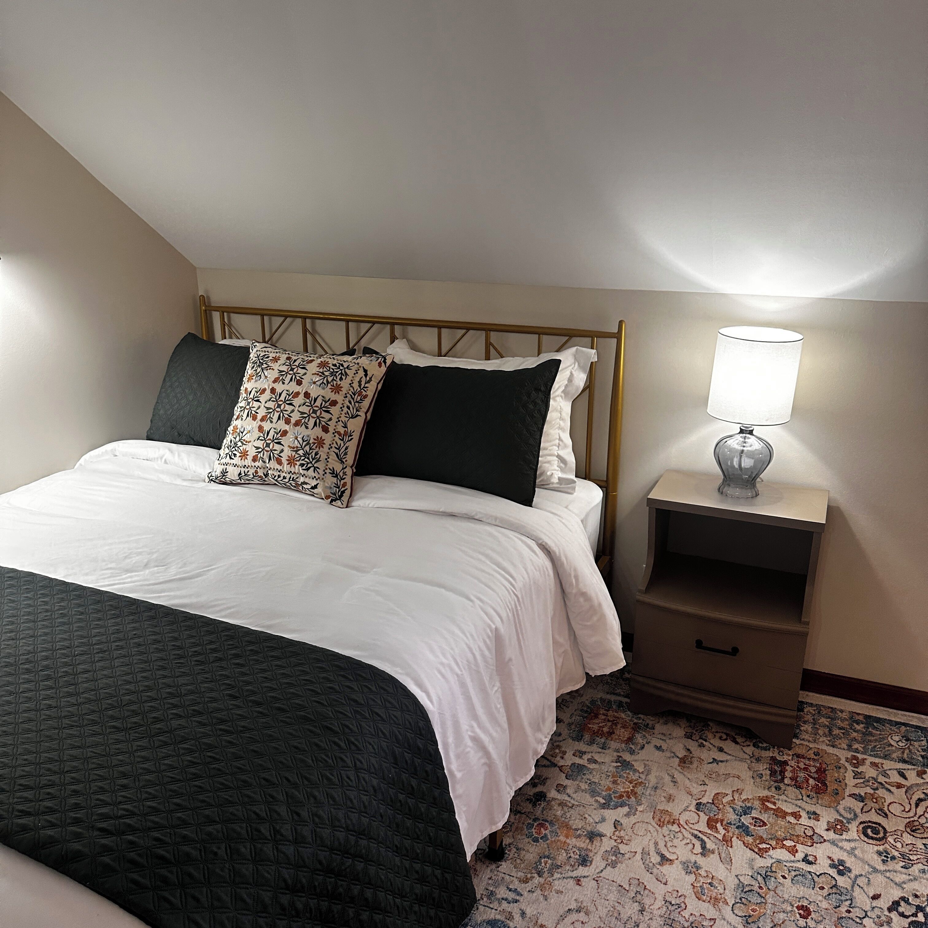 Ein Schlafzimmer mit einem Bett, Kissen, einem Nachttisch mit Lampe und einem gemusterten Teppich.