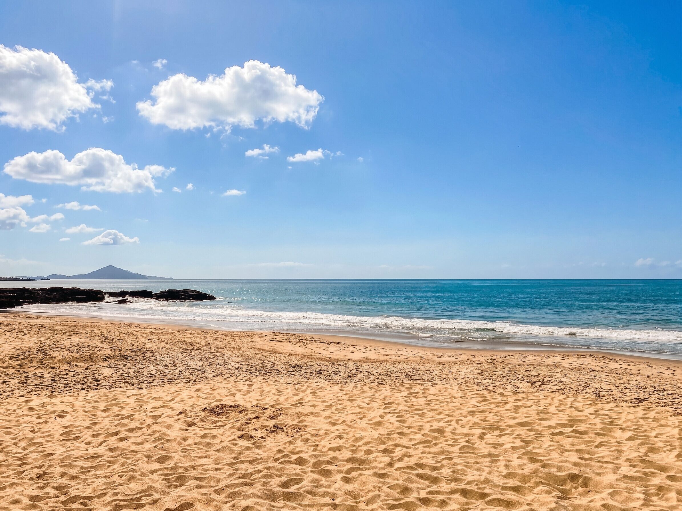 Uma praia com céu claro, ondas e uma montanha ao longe.
