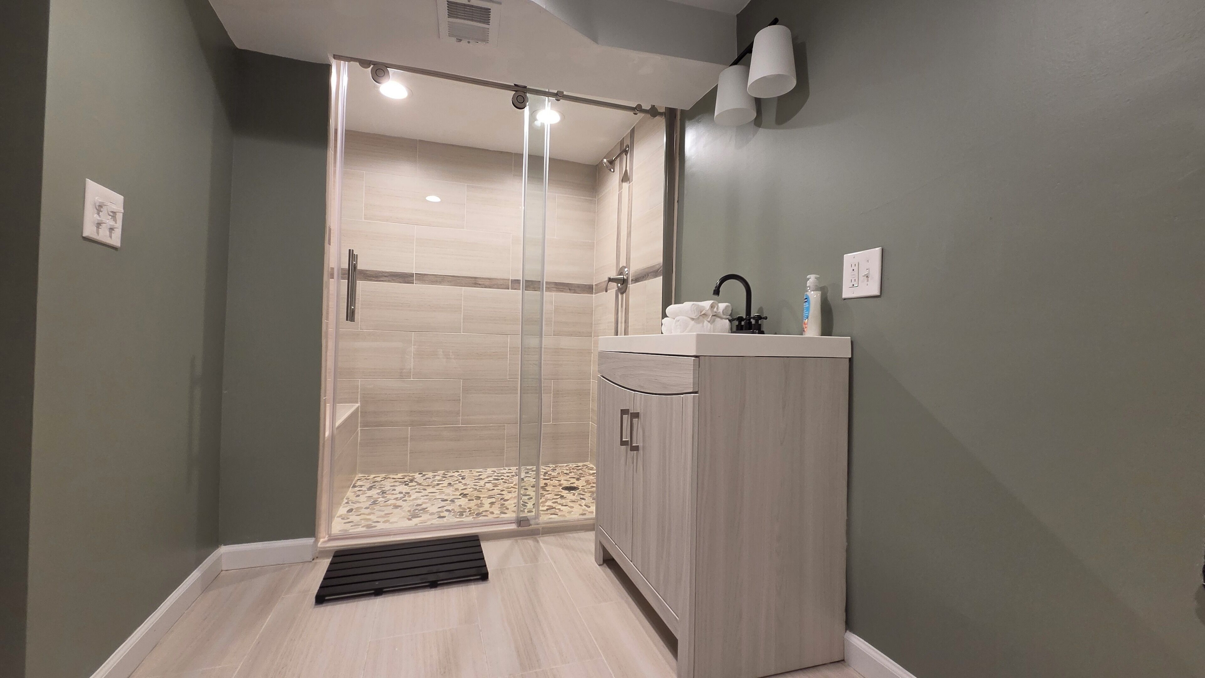 Une salle de bain moderne équipée d’une douche, d’un lavabo avec des articles de toilette et d’un luminaire.
