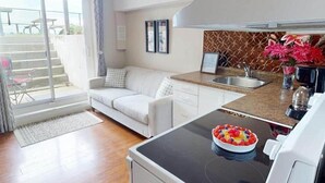 Een moderne keuken met een ingebouwde kookplaat, een zithoek met een witte bank en uitzicht op de omgeving via een schuifdeur.