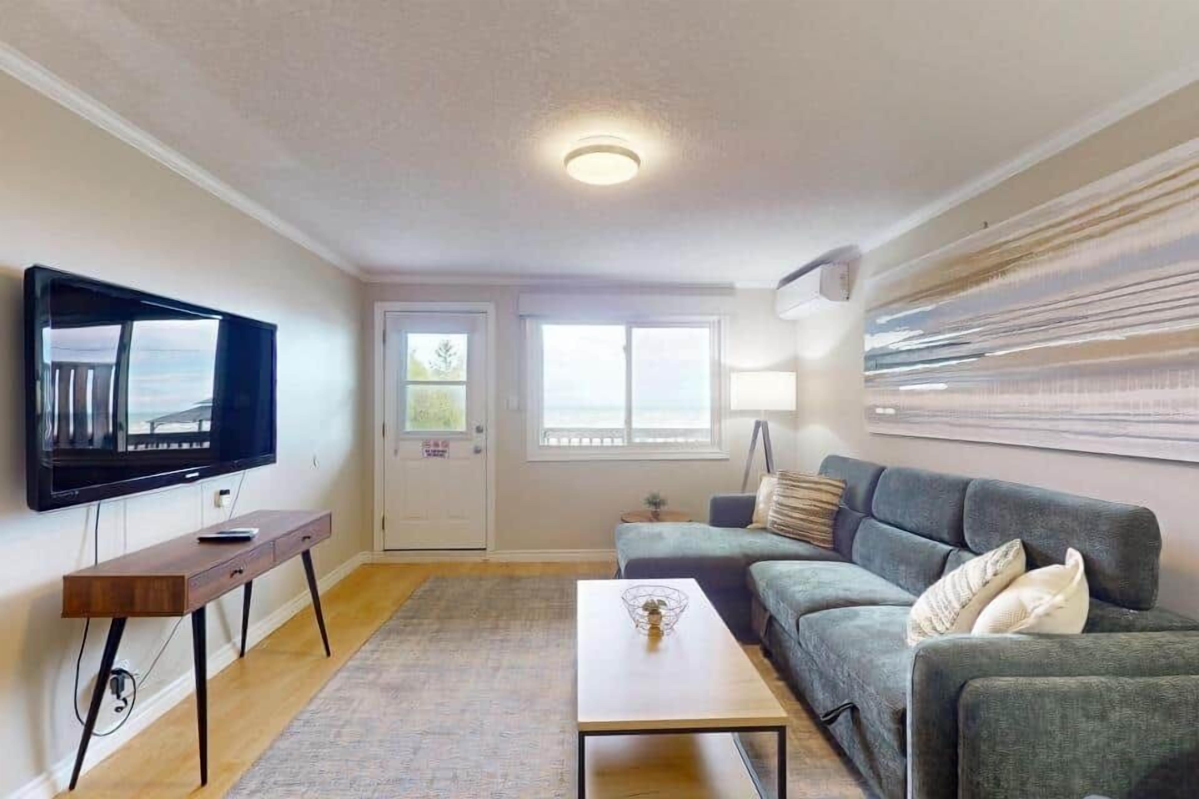 Ein Wohnzimmer mit Flachbildfernseher, Holztisch, grauem Sofa und großem Fenster.