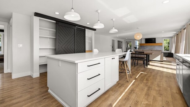 Een moderne keuken met witte kasten, een centraal eiland, hanglampen en een eethoek met een houten tafel en stoelen.