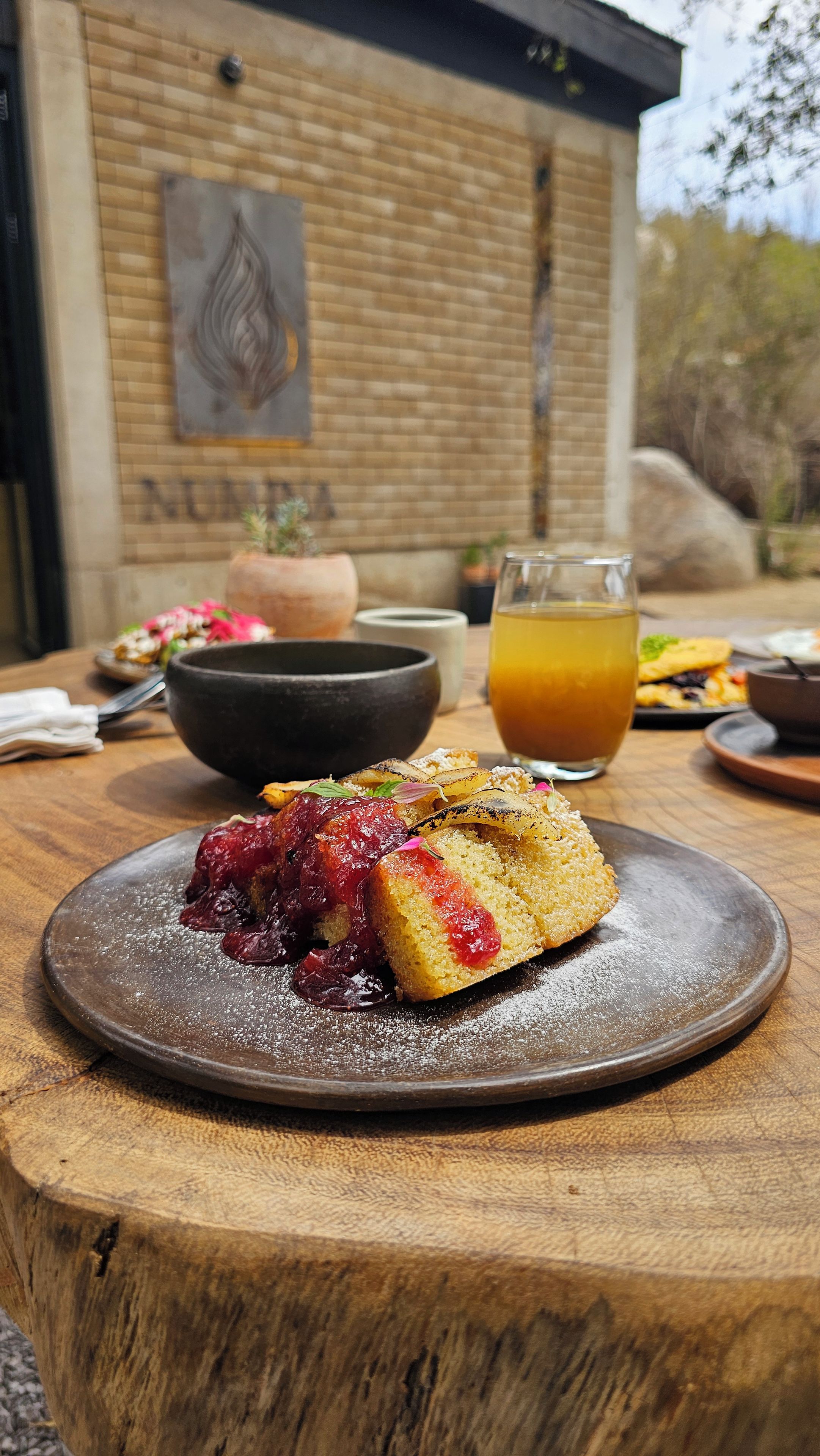 Un postre en un plato, una mesa de madera rústica, un vaso de jugo de naranja y un muro de ladrillo con una pintura de fondo.