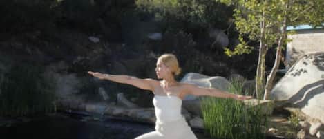 Una persona realizando una postura de yoga sobre una cubierta de madera con una colchoneta estampada, rodeada por un paisaje natural con rocas y vegetación.
