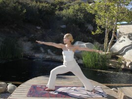 A person performing a yoga pose on a wooden deck with a patterned mat, surrounded by a natural landscape with rocks and vegetation.