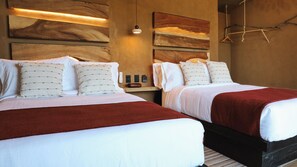 Habitación de hotel con dos camas, cada una con ropa de cama blanca y almohadas estampadas, una manta roja y un cabecero de madera.