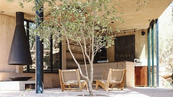 A covered outdoor seating area with two wicker chairs, a black chimney, and a stone wall.