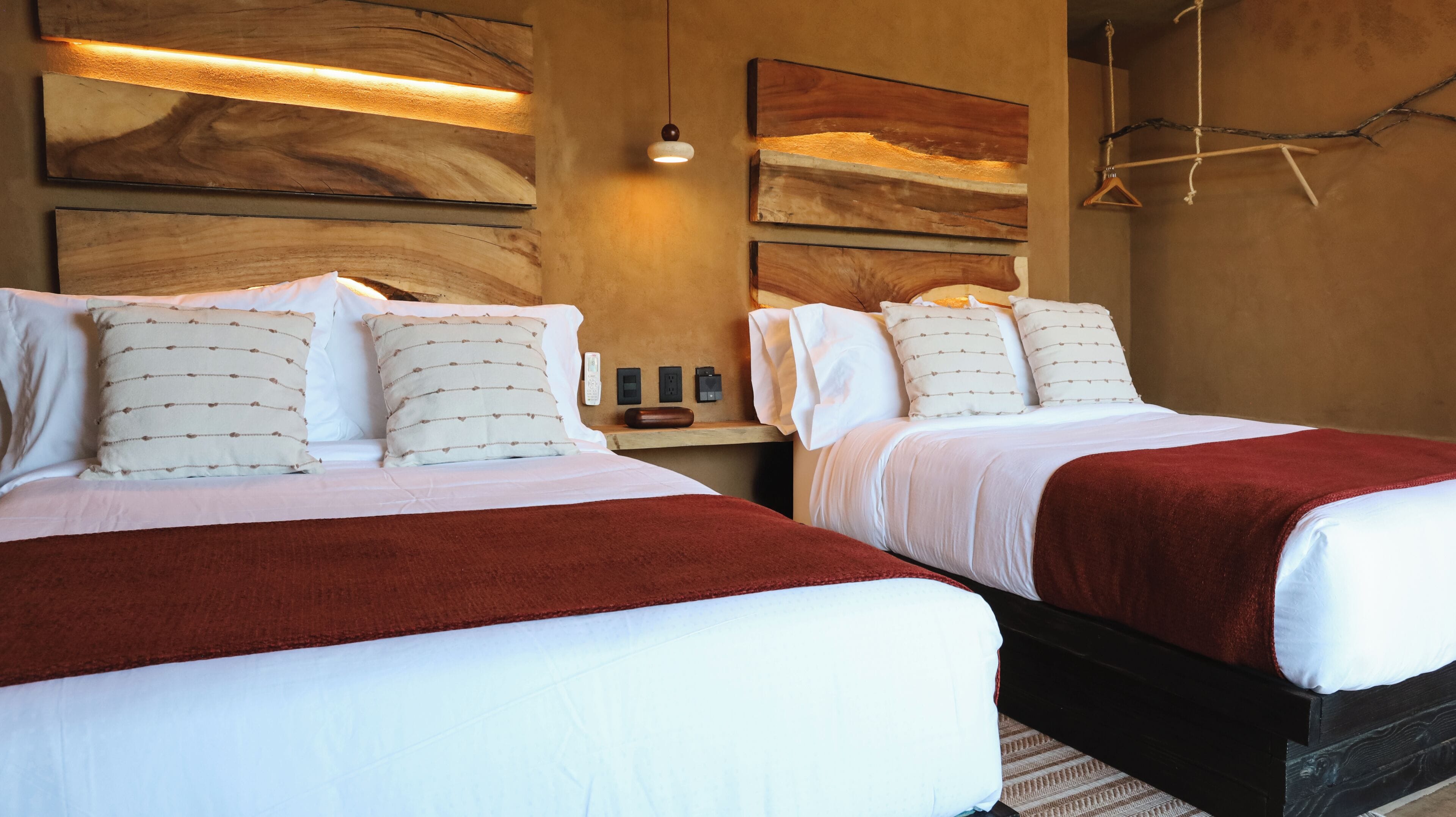 Habitación de hotel con dos camas, cada una con sábanas blancas y almohadas estampadas, cabecero de madera y una manta roja.