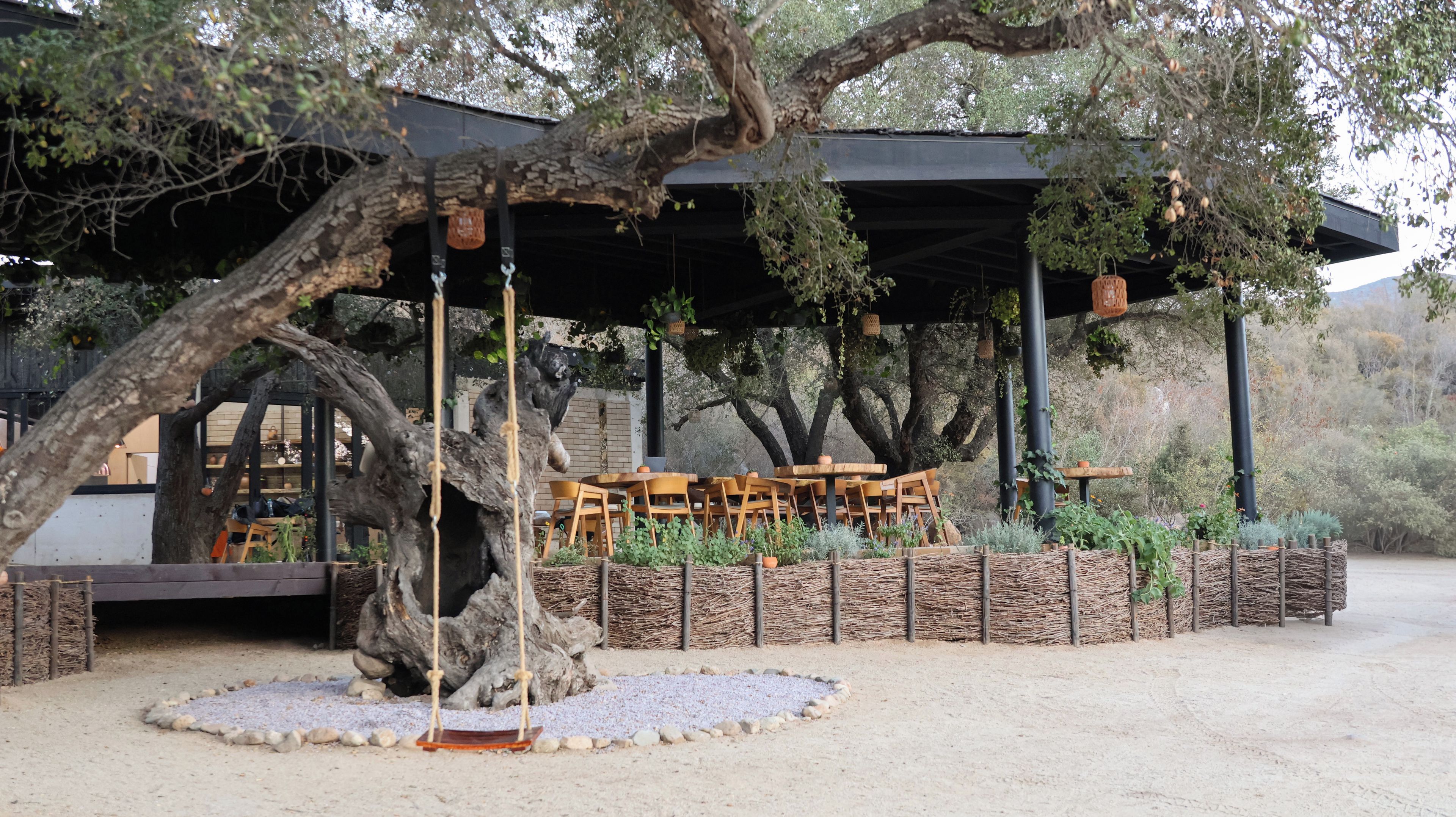 An outdoor seating area with wooden tables and chairs, surrounded by a rustic stone wall and greenery.