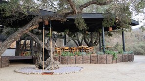 Un área de estar al aire libre con mesas y sillas de madera, rodeada por un muro de piedra rústico y vegetación.