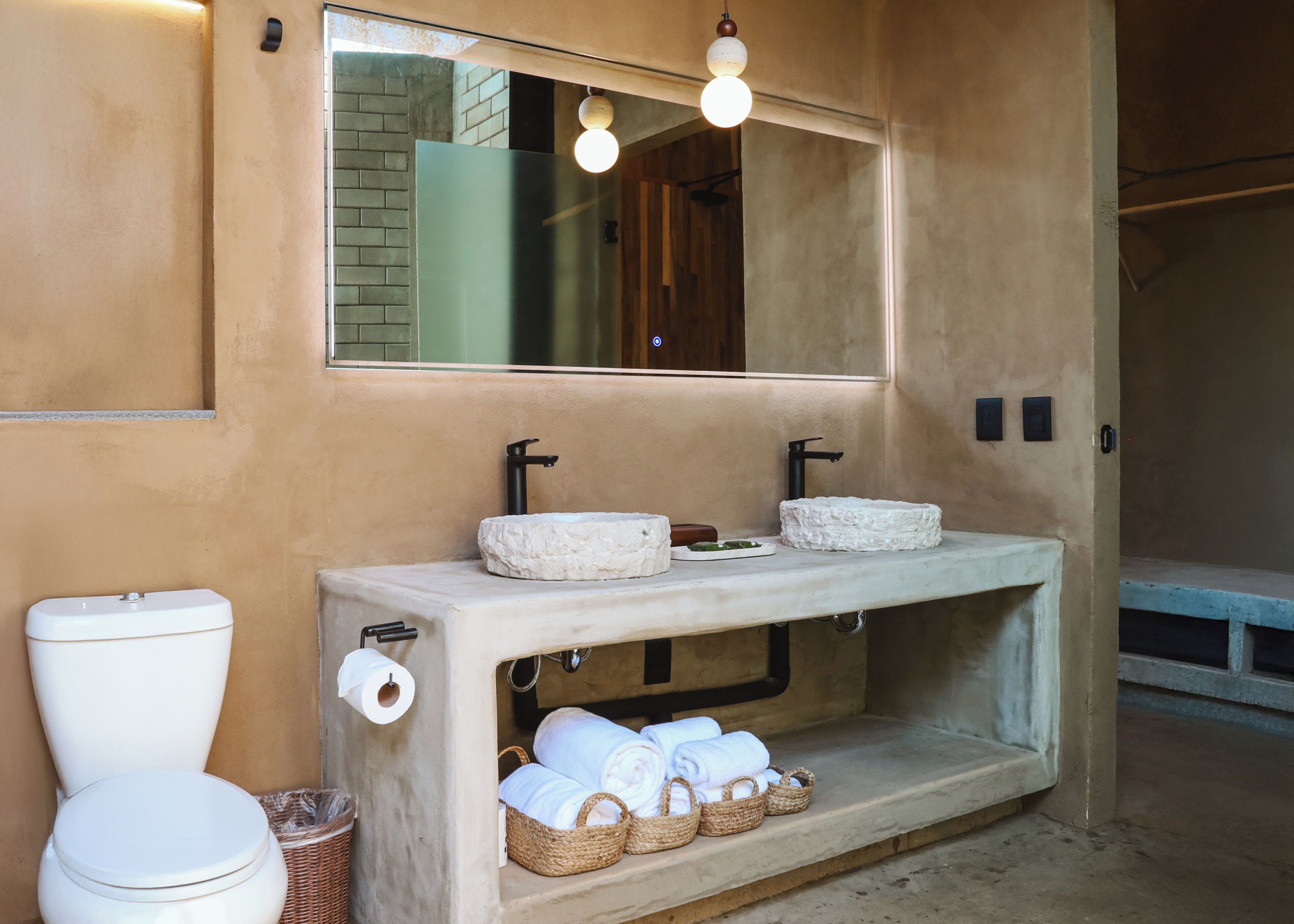 A bathroom with a toilet, two sinks, a mirror, and a shelf with towels and baskets.