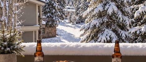 Una terraza de madera con dos botellas de cerveza, un bol de aperitivos y un menú, en un día soleado con árboles nevados y un telesilla al fondo.
