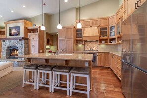 Eine moderne Küche mit Kamin aus Naturstein, Holzschränken, einem Edelstahlkühlschrank und einer Bar mit Hockern.