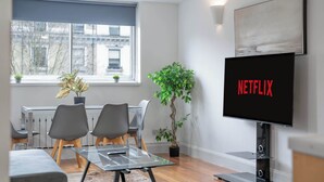Una sala moderna con una mesa de centro de vidrio, sillas grises, una planta en maceta, un televisor mostrando 'NETFLIX' y una ventana con persianas.