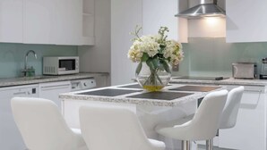 A modern kitchen with white cabinets, a stainless steel range hood, a microwave, and a dining area with a glass table and white chairs. - Luxurious Three Bedroom With Balcony (London)