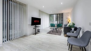 A modern living room with a flat-screen TV, a sofa, a dining table with chairs, and a large window with blinds. - Luxurious Three Bedroom With Balcony (London)