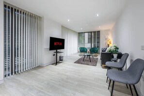 A modern living room with a flat-screen TV, a sofa, a dining table with chairs, and a large window with blinds. - Luxurious Three Bedroom With Balcony (London)