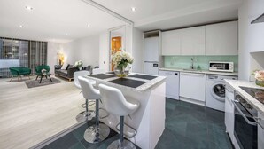 A modern kitchen with a central island, a dining area with bar stools, and a living room with a sofa and chairs. - Luxurious Three Bedroom With Balcony (London)