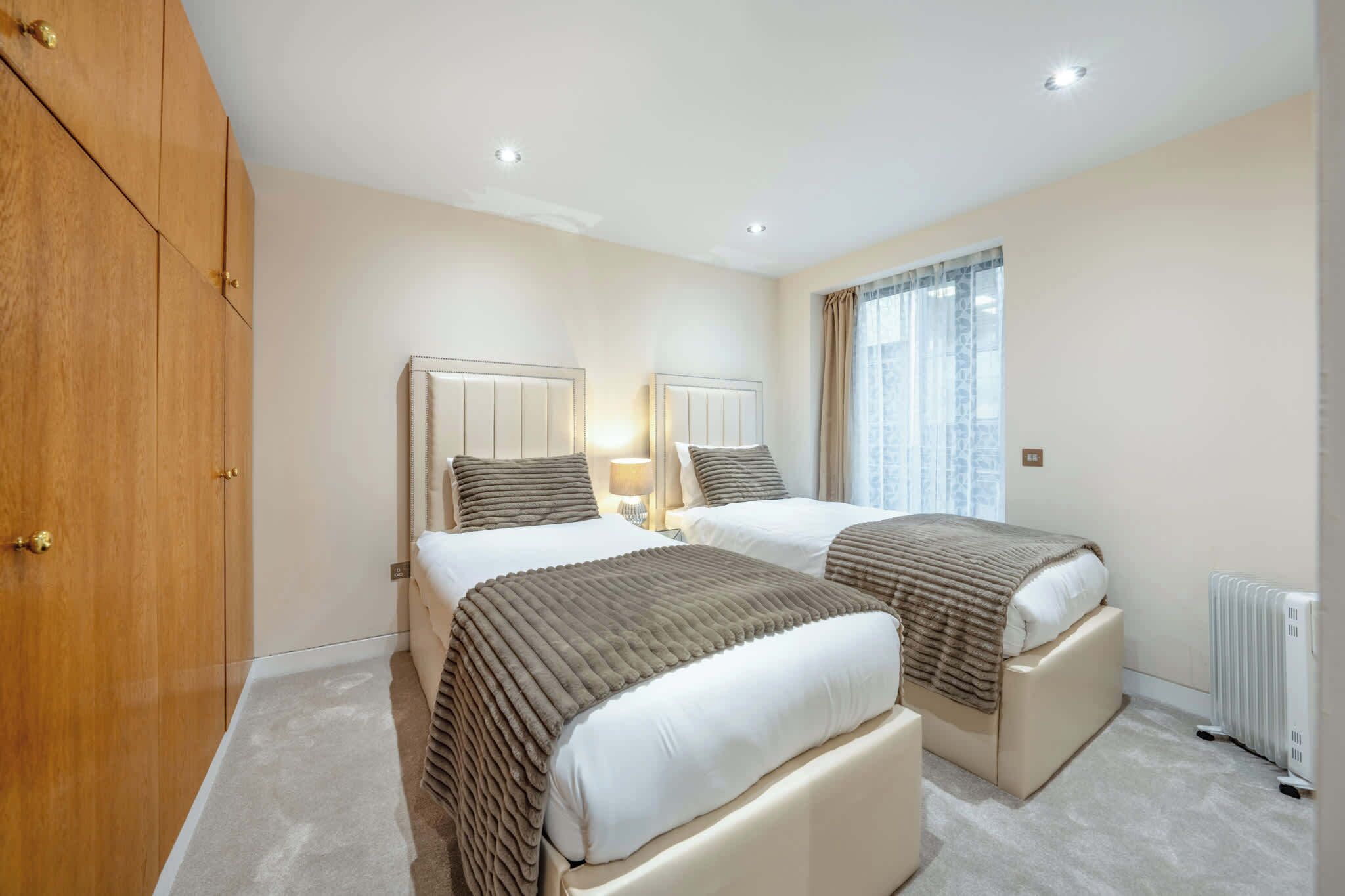 A hotel room with two beds, a wardrobe, a radiator, and a window with curtains.