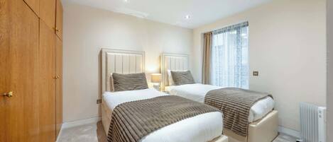 A hotel room with two beds, a wardrobe, a radiator, and a window with curtains.