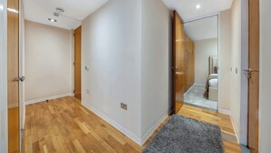 A hallway with wooden flooring, a grey rug, and a mirrored closet door reflecting a bedroom with a bed and a dresser. - Deluxe Two bedroom Apartment (London)