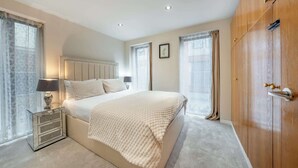 A bedroom with a bed, bedside table, lamp, curtains, and a wardrobe. - Deluxe Two bedroom Apartment (London)