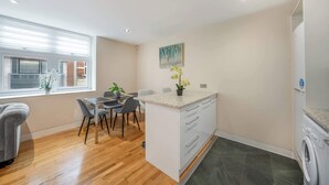 A modern kitchen with a dining area, a built-in washing machine, and a window with blinds. - Deluxe Two bedroom Apartment (London)