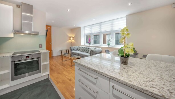 A modern kitchen with a built-in oven, a dining area with a sofa and table, and a large window with natural light. - Deluxe Two bedroom Apartment (London)