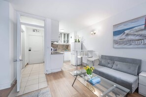 Un salon moderne avec un canapé gris, une table basse en verre ornée de fleurs, une cuisine en arrière-plan et un tableau représentant un phare accroché au mur.