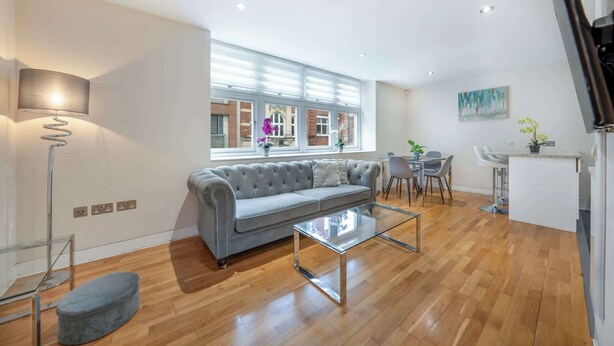 Uma sala de estar moderna com um sofá cinzento, uma mesa de centro de vidro, um abajur e um espaço de jantar com uma mesa e cadeiras brancas.