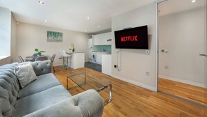 Uma sala de estar moderna com um sofá cinzento, uma mesa de centro de vidro, uma TV de tela plana na parede e uma área de cozinha ao fundo.