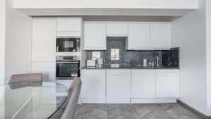 A compact kitchen with white cabinetry, a glass table, and a herringbone-patterned floor.
