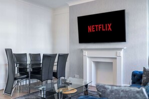 A modern living room with a glass table, black chairs, a fireplace, and a TV displaying 'NETFLIX'. - Luxurious Two Bedroom Two Bathroom (London)