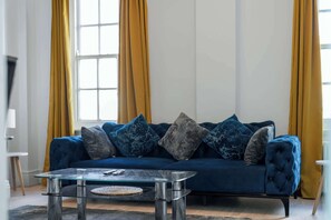 A blue sofa with multiple cushions, a glass coffee table, and yellow curtains. - Luxurious Two Bedroom Two Bathroom (London)