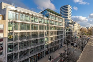 A multi-story building with large windows, a curved structure to the right, and a street with cars and a tram. - Luxurious Two Bedroom Two Bathroom (London)