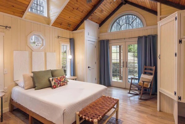Ein Schlafzimmer mit einem großen Bett, einer Holzbank, einem Schaukelstuhl und Blick ins Freie durch eine Tür.