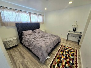 Ein Schlafzimmer mit einem Bett, Nachttischen, einem kleinen Schreibtisch, einem Stuhl, einem gemusterten Teppich und einem Fenster mit transparenten Vorhängen.